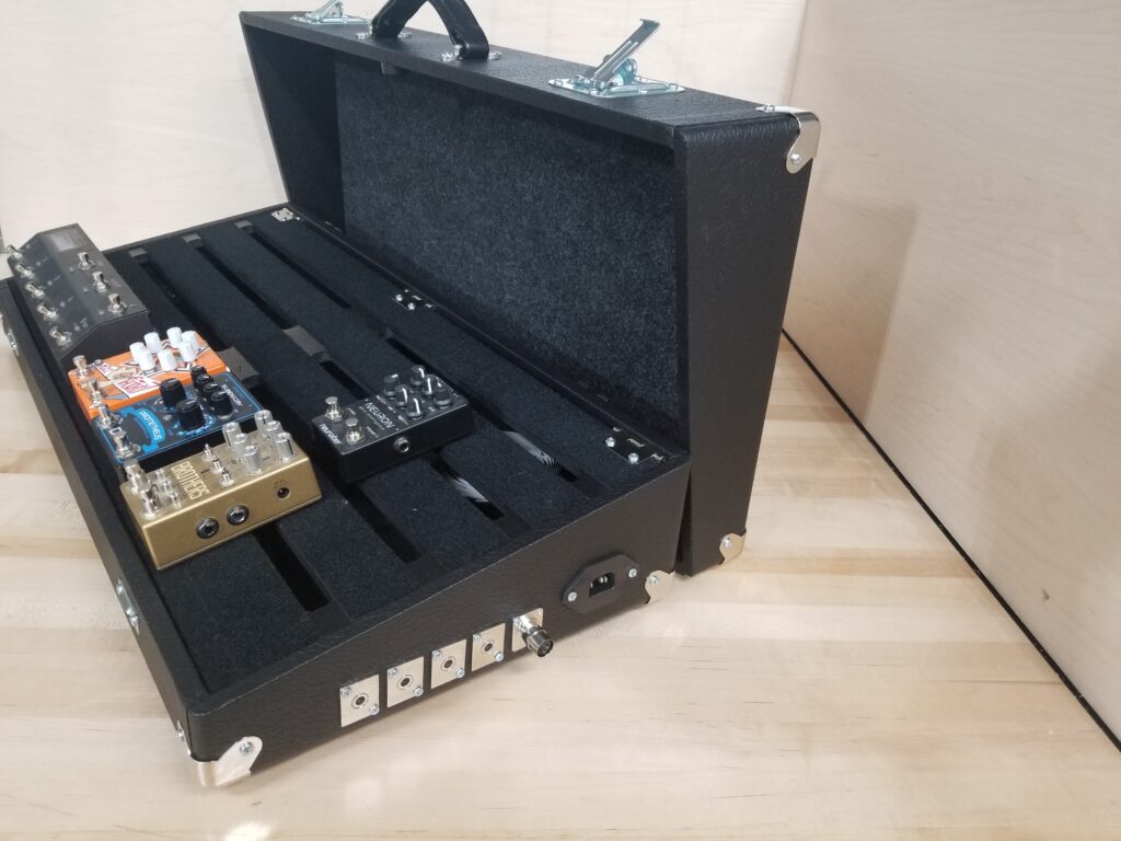 A Pedal Pad pedal board with 5 pedals on the pedal deck. The side of the board has 5 audio ins/outs and an IEC inlet for integrated pedal board power solutions