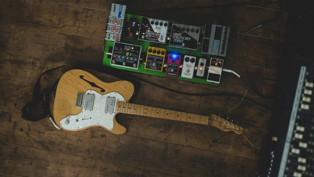 A guitar next to a pedal board on the ground with a power supply in the top corner